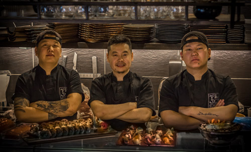 Tre män med svarta kockkläder står i ett restaurangkök med armarna i kors